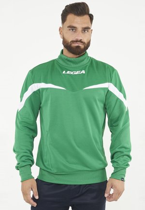 Muž s vousy v zelené sportovní bundě s bílými proužky a logem "LEGEA", stojící na bílém pozadí.