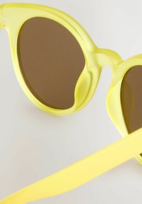 Sárga napszemüveg áttetsző műanyag kerettel és barna lencsékkel. A design lekerekített szélekkel és vékony, lapos templivel rendelkezik.