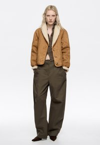 Chaqueta de ante marrón con un cuello de borrego crema, combinada con pantalones de pierna ancha verde oliva y zapatos marrones de punta.