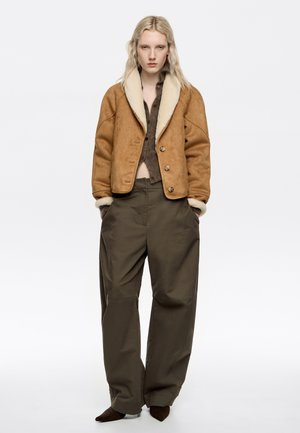 Chaqueta de ante marrón con un cuello de borrego crema, combinada con pantalones de pierna ancha verde oliva y zapatos marrones de punta.