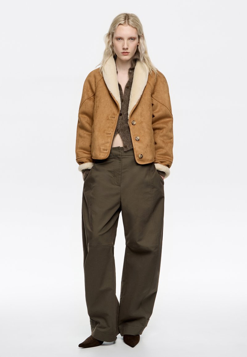 Chaqueta de ante marrón con un cuello de borrego crema, combinada con pantalones de pierna ancha verde oliva y zapatos marrones de punta.