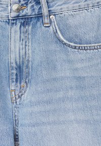 Hellblaue Jeans aus Denim im Nahaufnahme, die den vorderen Knopf, die Gürtelschlaufen und die teilweise sichtbare Vordertasche mit Stickdetails zeigt.