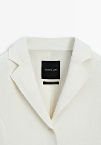 Blazer w kolorze off-white z klapami w kształcie notcha i pojedynczym guzikiem, z widoczną czarną metką wewnętrzną z logo marki oraz napisem "Wyprodukowano w Portugalii".