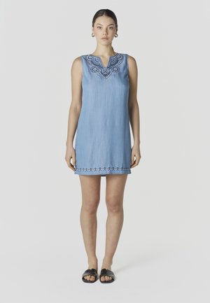 Mujer de pie con un vestido sin mangas azul claro con bordados azul oscuro en el escote y el dobladillo, sandalias negras abiertas en los dedos y pendientes de aro.