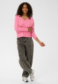 Rozā v-veida adīta džemperis ar brīvu piegriezumu un ribinātām aprocēm, apvienots ar augsta vidukļa melnajām džinsu biksēm un pelēkajām sporta apaviem.