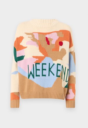 Nainen pukeutunut värikkääseen "WEEKEND"-neuleeseen, vaaleansiniset leveälahkeiset farkut, mustat terävät kengät, pitää valkoista punottua käsilaukkua.