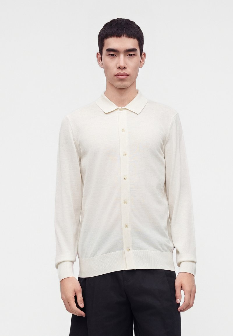 Jeune homme portant une chemise blanche à manches longues boutonnée et un pantalon noir, debout sur un fond blanc uni.