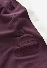 Temno rdeč športni material z belo kontrastno obrobo; vsebuje vtisnjene detajle "by" in številko "0" z teksturiranim zaključkom. Mehka in gladka tekstura.