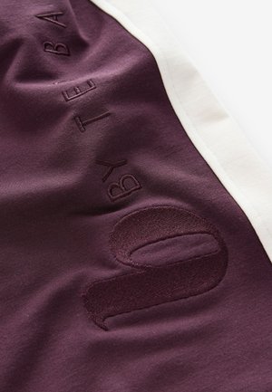 Tessuto atletico color granata con rifiniture bianche a contrasto; presenta dettagli in rilievo con la scritta "by" e il numero "0" con finitura testurizzata. Tessuto morbido e liscio.