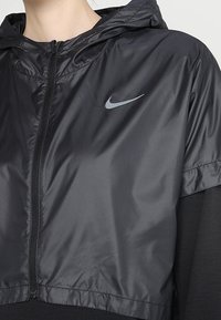 Veste courte noire en matériau léger et brillant. Dispose d'une capuche, d'une fermeture éclair, et d'un petit logo Nike gris sur la poitrine.