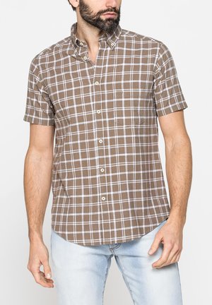 Uomo con la barba che indossa una camicia marrone a quadri a maniche corte e jeans azzurri, in piedi davanti a uno sfondo semplice.