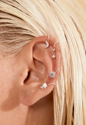 Ucho s piatimi striebornými náušnicami, vrátane polmesiaca, malej perly, slnečného lúča a kvetinových motívov na pozadí blond vlasov.