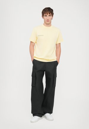 Yellow short-sleeve t-shirt with text graphic, paired with wide black cargo pants featuring side pockets and white sneakers.