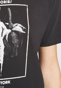 Black cotton T-shirt featuring a graphic print of a pepper and text. The design includes a rectangular border and is fitted at the sleeve.