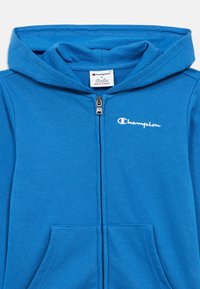 Μπλε φούτερ με φερμουάρ κατασκευασμένο από υφή υφάσματος, με εμπρόσθιο φερμουάρ, δύο ανοίγματα για τσέπες και ένα μικρό λευκό λογότυπο "Champion" στο στήθος.