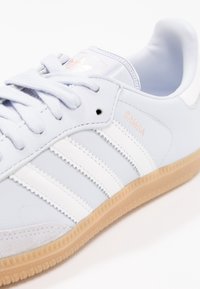 Světlounce modrá kožená teniska se světlými bílými pruhy, texturovanými semišovými akcenty a podrážkou z gumové pryže. Logo "SAMBA" vyražené v růžově zlaté barvě.