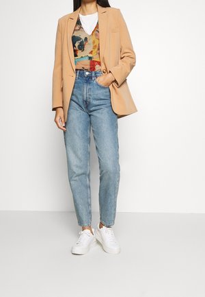 Femme portant un blazer beige, un gilet à motifs par-dessus une chemise blanche, un jean bleu clair taille haute et des baskets blanches, debout avec une main dans la poche.