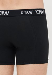 Ingrandimento di una persona che indossa boxer neri con una cintura nera che mostra il logo bianco "ICIW".