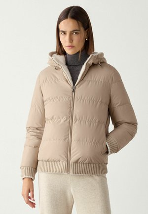 Frau trägt eine beige Steppjacke mit Kapuze, gerippten Bündchen und Saum, kombiniert mit hellen Hosen und einem dunklen Rollkragenpullover.