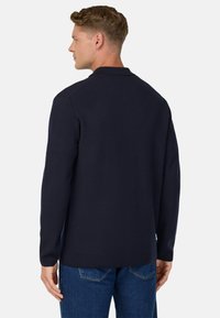 Jūras zila garām piedurknēm blūze ar apkakli un minimālu tekstūru. Iezīmē taisnās griezuma un tiek valkāta ar zilām džinsu biksēm.