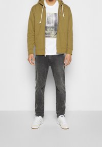 Felpa con zip color verde oliva, t-shirt bianca con stampa di un albero, jeans grigi e sneakers bianche con righe nere. Sfondo semplice.