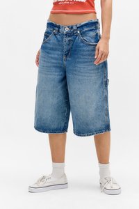 Pantalones cortos de mezclilla azul claro con un corte holgado, de longitud media por encima del muslo, cinco bolsillos y cierre de botón. Llevados con zapatillas blancas y calcetines acanalados.