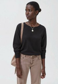 Schwarzer Rundhalsausschnitt-Pullover mit langen Ärmeln, kombiniert mit hellbraunen Jeans. Das Model trägt eine beige Umhängetasche. Eine silberne Halskette ist sichtbar.
