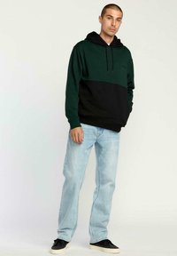 Joven con una sudadera verde y negra, jeans de color azul claro y zapatillas negras, de pie contra un fondo claro.