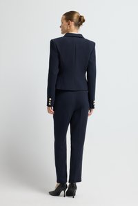 Granatowy garnitur dwuczęściowy z marynarką o zdefiniowanej konstrukcji i dopasowanymi spodniami. Marynarka ma mankiety z złotymi guzikami, gładką teksturę oraz dopasowaną sylwetkę.