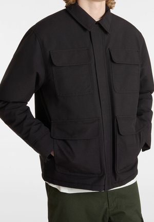 Person trägt eine schwarze Jacke mit vier Klappentaschen vorne, kombiniert mit olivgrünen Hosen und einem weißen Hemd darunter.