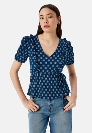 Haut bleu marine avec un décolleté en V, des manches courtes à volants et un accent à nouer à la taille. Présente un motif géométrique blanc sur un tissu texturé.