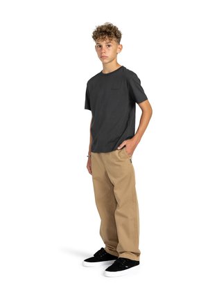 Boy wearing a short-sleeve dark gray t-shirt and tan loose-fit pants, with black sneakers, standing in a relaxed pose against a white background.