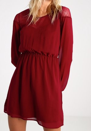 Femme portant une robe rouge profond à manches longues, longueur genou, taille cintrée et panneaux en tissu transparent sur les épaules et les manches.