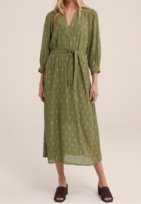 Grön långärmad klänning med v-ringning, bältad midja och en flödande silhuett. Har ett texturerat tyg med lätta mönster genomgående.