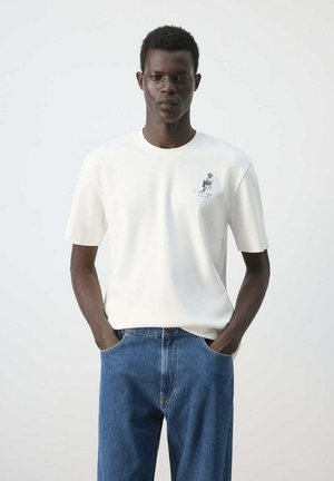 Hombre que lleva una camiseta blanca de manga corta con un pequeño gráfico en el pecho y vaqueros azules, de pie con las manos en los bolsillos.