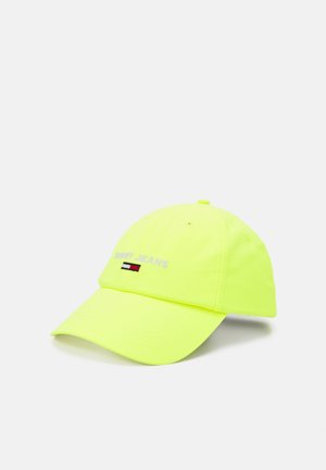 Casquette de baseball jaune vif avec le texte « Tommy Jeans » et un logo brodé rouge, blanc et noir sur le devant, reposant sur une surface blanche.