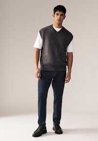 Svetari s V-izrezom bez rukava u tamnosivom triku preko bele majice, uparen sa tamnoplavim farmerkama i crnim čizmama na pertlanje. Glatka tekstura.