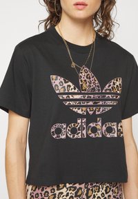 Frau trägt ein schwarzes, kurzes Adidas-T-Shirt mit Leopardenmuster-Logo, goldene, geschichtete Halsketten und passende Hosen im Leopardenmuster.