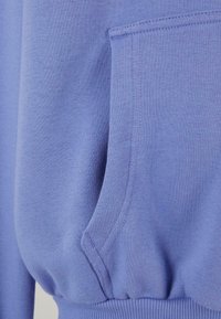 Sweatshirt de culoare lavandă deschisă cu buzunar tip cangur, mansete și talie ribbate. Material moale cu textură netedă și detalii de cusătură vizibile.