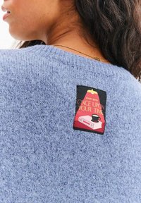 Jasnoniebieski dzianinowy sweter o strukturalnej fakturze, z czarną metką przedstawiającą książkę i filiżankę kawy oraz napisem "START WITH ONCE UPON YOUR TIME."