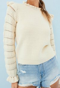 Femme portant un pull en maille crème avec des épaules et des poignets à volants, assorti à un short en jean bleu clair déchiré, sur un fond bleu pâle.