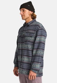 Chemise en flanelle avec un motif rayé vert et marron, devant à boutons, deux poches poitrine et manches longues. Accompagnée d'un bonnet noir.