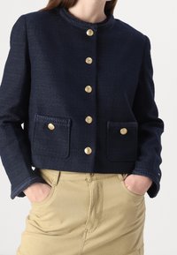 Navy blue textured jacket with round neckline, five gold buttons, and two braided pocket accents. Worn over beige pants.