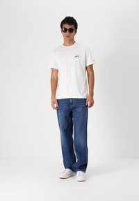 Witte katoenen t-shirt met een klein zwart logo, gecombineerd met blauwe spijkerbroek en witte sneakers. Het model draagt zwarte zonnebril.