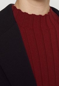 Gros plan sur une personne portant un pull côtelé rouge foncé avec un col festonné sous un blazer noir.