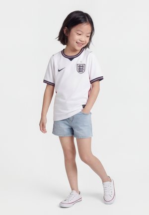 Jong meisje dat lacht, gekleed in een wit Engels voetbalshirt, lichtblauwe denim korte broek en witte sneakers, staand met één hand in de zak.