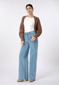 Καφέ πλεκτή ζακέτα πάνω από λευκό εφαρμοστό τοπ, συνδυασμένη με ψηλόμεσα ανοιχτό μπλε παντελόνια με φαρδιά μίντια και λεπτομέρειες με κουμπιά. Μπεζ παπούτσια με τακούνι.