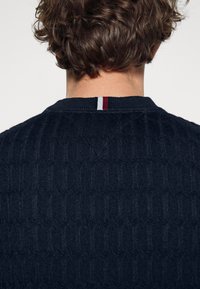 Marineblauwe gebreide trui met een gestructureerd patroon, geribde halslijn en een klein driemaal kleuraccent bij de achterkraag.