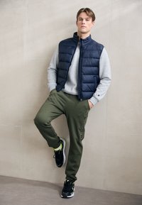 Marineblauer Steppweste über einem grauen Sweatshirt, kombiniert mit olivgrünen Jogginghosen und schwarzen Sneakers mit neonfarbenen Akzenten. Texturierte Materialien sichtbar.