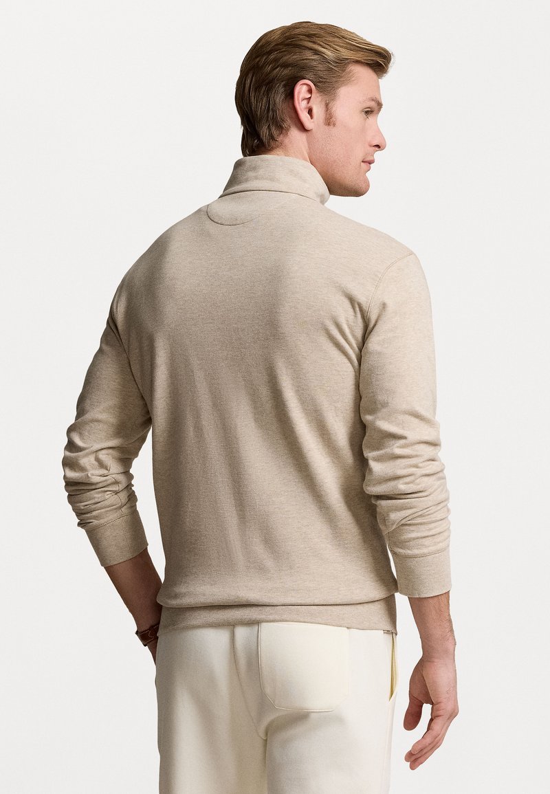 Pullover beige con colletto alto, polsini a coste e una superficie liscia. Il capo ha una vestibilità rilassata ed è realizzato in materiale morbido.